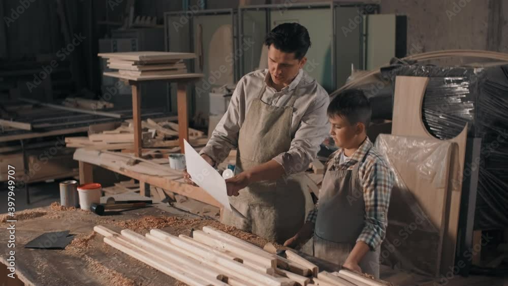 Medium shot of father and son spending time together in carpentry workshop looking at blueprint of diy furniture made of wood Medium shot of father and son spending time together in carpentry workshop looking at blueprint of diy furniture made of wood