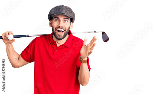 Young handsome man with beard playing golf holding club and ball celebrating victory with happy smile and winner expression with raised hands