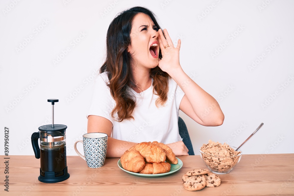 Young beautiful brunette woman sitting on the table eating breakfast in the morning shouting and screaming loud to side with hand on mouth. communication concept.