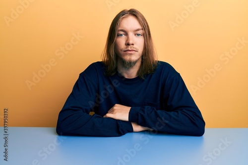 Handsome caucasian man with long hair wearing casual clothes sitting on the table relaxed with serious expression on face. simple and natural looking at the camera. © Krakenimages.com