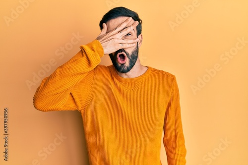 Young hispanic man wearing casual clothes peeking in shock covering face and eyes with hand, looking through fingers with embarrassed expression.
