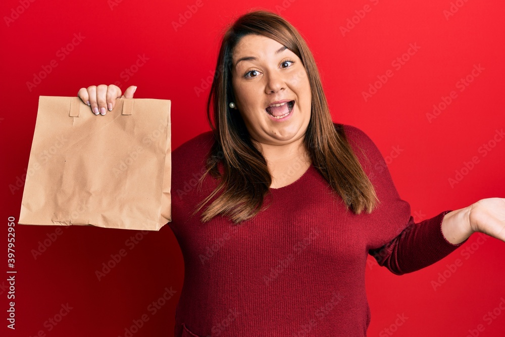 Beautiful brunette plus size woman holding take away paper bag celebrating achievement with happy smile and winner expression with raised hand Beautiful brunette plus size woman holding take away paper bag celebrating achievement with happy smile and winner expression with raised hand