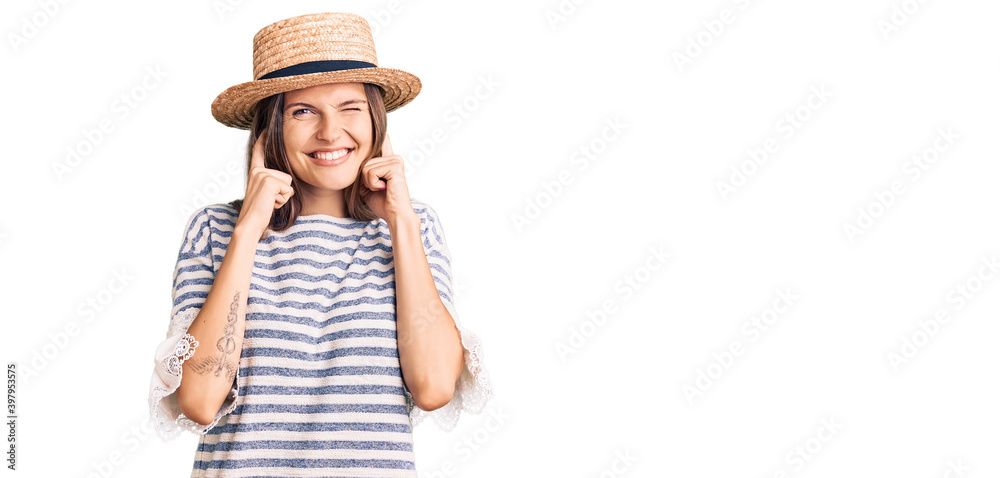 Beautiful caucasian woman wearing summer hat covering ears with fingers with annoyed expression for the noise of loud music. deaf concept. Beautiful caucasian woman wearing summer hat covering ears with fingers with annoyed expression for the noise of loud music. deaf concept.