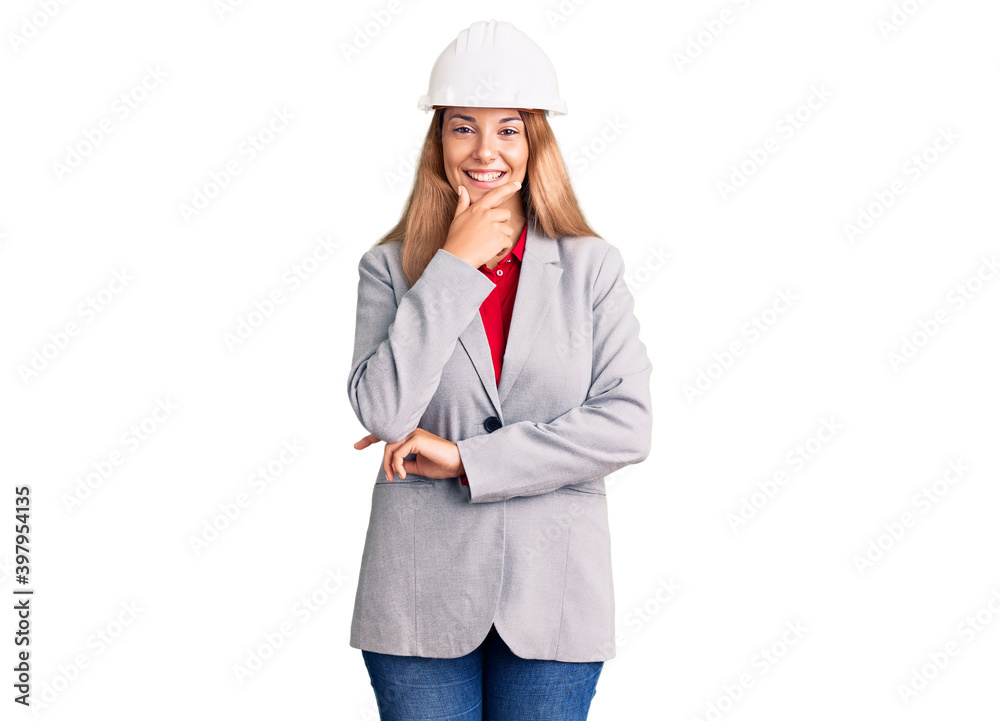 Beautiful young woman wearing architect hardhat looking confident at the camera smiling with crossed arms and hand raised on chin. thinking positive.