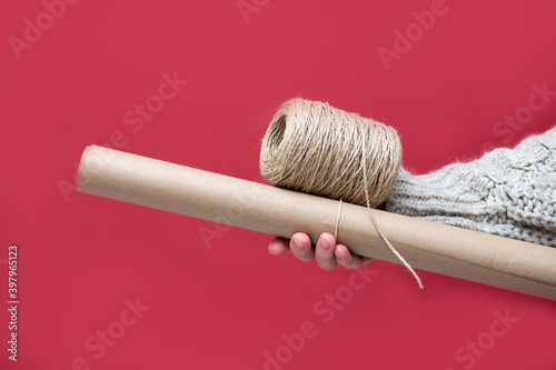 Hand holding a roll of Kraft paper and a reel of rope on a red background, close-up. Gift wrapping with your own hands in a retro style concept