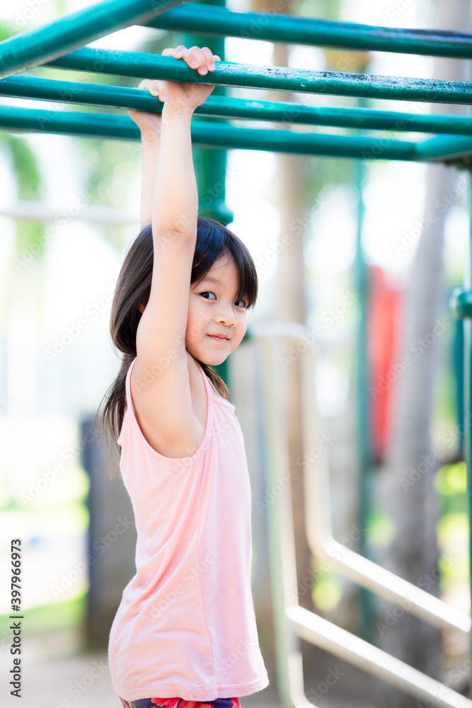 Fototapeta premium Active cute girl playing hanging outdoors. 5 years kid climbing bar at playground near by the house. Happy child enjoying activity in a climbing adventure park on summer day. Children sweet smiles.