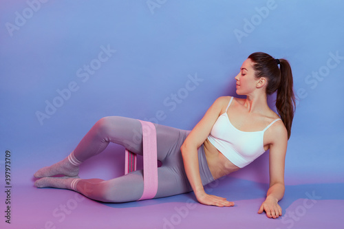 Health and sports, winsome lady wearing white top and gray leggins training inside leg while lying on floor, used fitness rubber band, young girl being in fit, working out with equipment.