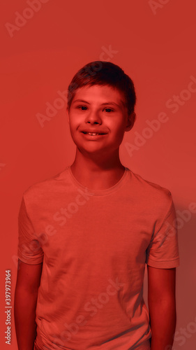 Portrait of joyful teenaged disabled boy with Down syndrome smiling at camera while posing isolated over red light background
