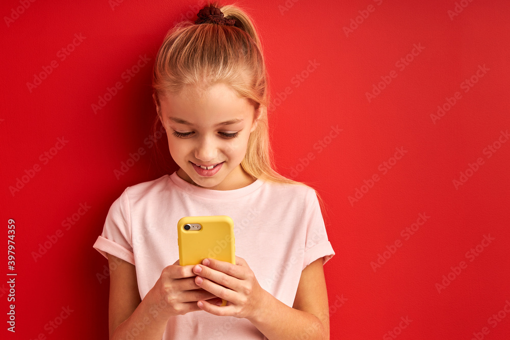 child girl watching videos on smartphone, isolated over red background in studio. little girl with ponytail hold yellow mobile phone in hands, smile looking at screen child girl watching videos on smartphone, isolated over red background in studio. little girl with ponytail hold yellow mobile phone in hands, smile looking at screen