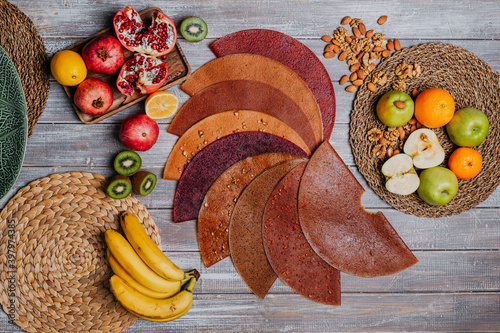 Many colorful fruit leathers with fresh fruits on the wooden table. Round fruit leather. Healthy food. Apples, bananas, nuts, lime, kiwifruits, pomegranate top view.