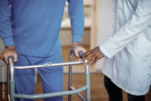 Unrecognizable elderly man being assisted to walk with mobility walker at nursing home.