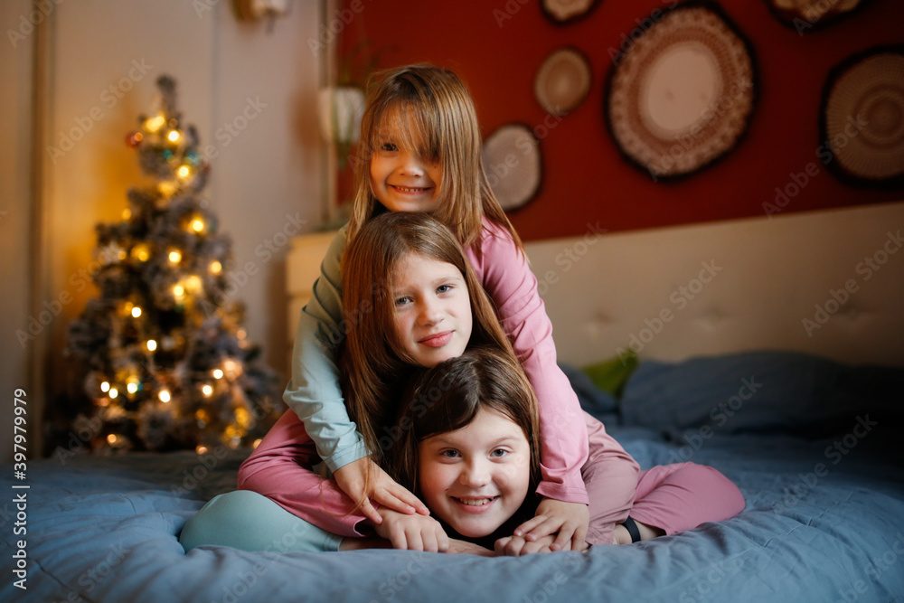 three Caucasian girls children in pajamas rage and hug on bed in bedroom near of Christmas tree. Concept of childhood and anticipation of the new year, cozy home children's photos three Caucasian girls children in pajamas rage and hug on bed in bedroom near of Christmas tree. Concept of childhood and anticipation of the new year, cozy home children's photos