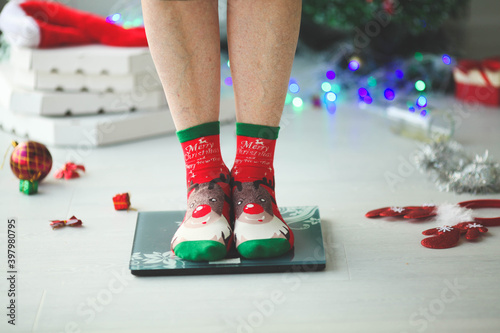 Close-up of adult woman legs in socks with deer stand on scales measure weight.  After the holiday Christmas, pizza boxes and New Year's decorations, bottles are lying on the floor.