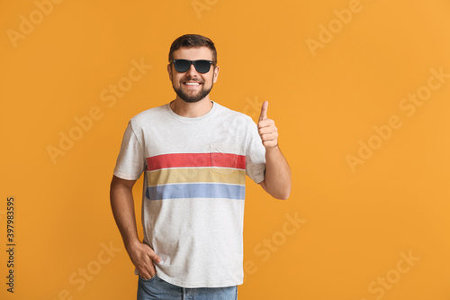 Handsome man with stylish sunglasses showing thumb-up gesture on color background