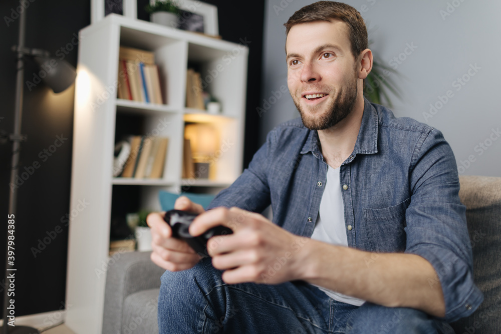 Excited young man relaxing on grey couch and using joystick for playing video games. Bearded guy having fun during free tim at home. Excited young man relaxing on grey couch and using joystick for playing video games. Bearded guy having fun during free tim at home.