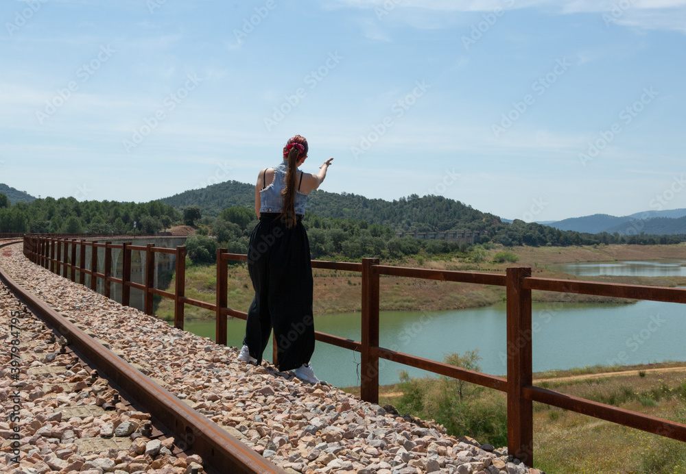 Mujer contenta de espaldas, sobre una vía de tren abandonada en verano., disfrutando de vistas de lago, en actitud divertida y relajada. Estilo de vida.