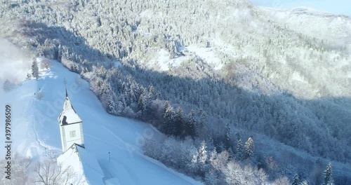 Aerial drone flying over famous Jamnik church, Slovenia. Landscape covered with white snow. Fairy tale like nature and small catholic church on hilltop. Dynamic moving, parallax photo