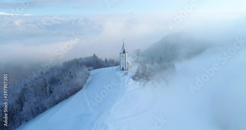 Aerial drone flying over famous Jamnik church, Slovenia. Landscape covered with white snow. Fairy tale like nature and small catholic church on hilltop. Alps mountains in the distance