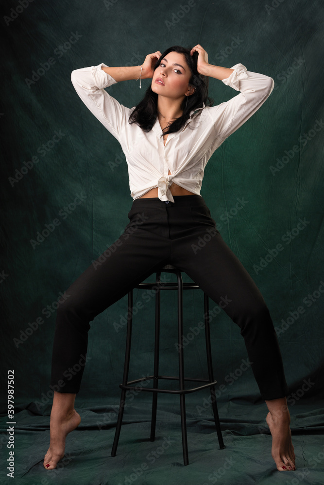 a beautiful brunette girl in black trousers and a white shirt sits basik on a high stool. woven background backlit in green. a beautiful brunette girl in black trousers and a white shirt sits basik on a high stool. woven background backlit in green.