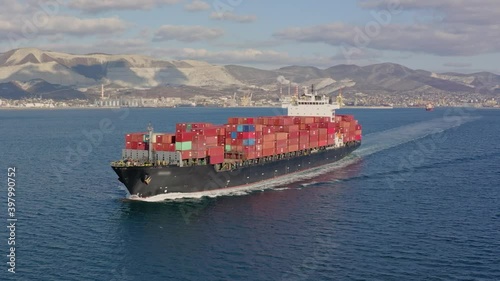 Aerial view of container ship sailing across the sea departing from the port on sunny warm summer day. Concept of sea delivery, logistics. Drone high quality footage