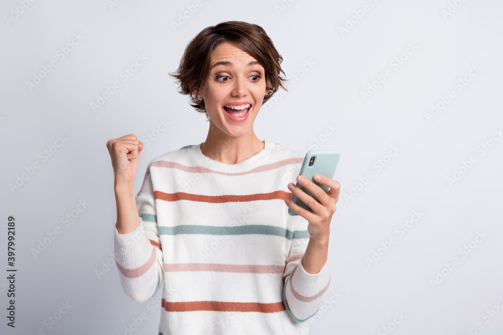Photo of lucky young woman dressed striped pullover looking holding modern device arm fist isolated grey color background Photo of lucky young woman dressed striped pullover looking holding modern device arm fist isolated grey color background