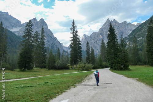 A woman with a big hiking backpack hiking in the Italian Dolomites. There are high and sharp mountain peaks in front of her. She walks on a wide, gravelled road surrounded by a few trees. Remedy photo