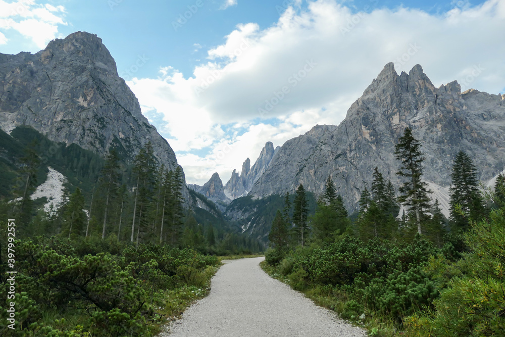 A gravel road through a forest, leading to high and sharp Italian Dolomites. The road is very curvy. There are thick clouds above the mountain peaks. Idyllic landscape. Isolated and remote place