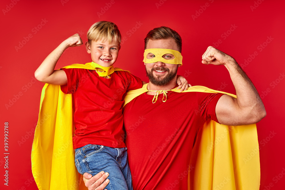 brave and strong father with son in superhero costume, they show arm muscles at camera, wearing eyewear, isolated over red background