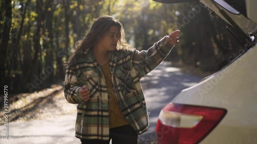 Happy young woman in checkered jacket opens trunk of white automobile to take map and thermocup on empty rural road in picturesque autumn forest