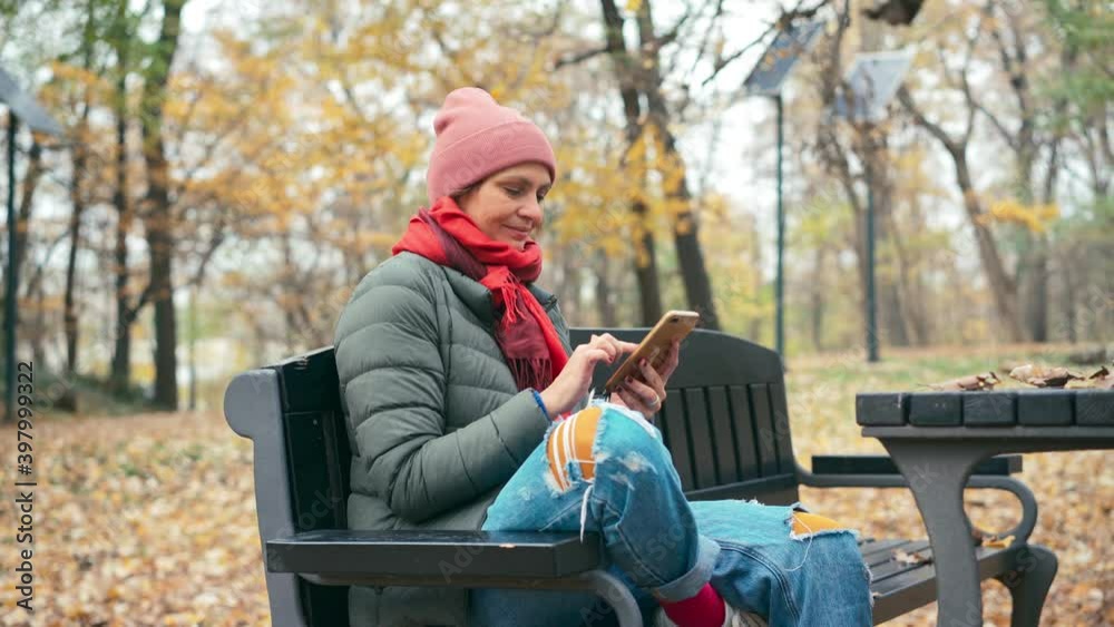 A young woman in a jacket and a hat using her smartphone while sitting on the bench in the autumn park. A young woman in a jacket and a hat using her smartphone while sitting on the bench in the autumn park.