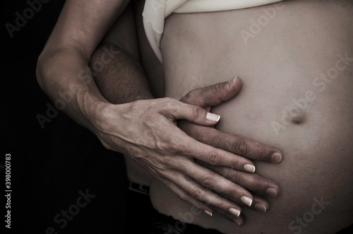 The belly of a pregnant woman being hugged by a man’s hand and her own hand. You can see the belly, the hands and the belly button. You can also see a part of a white t-shirt.
