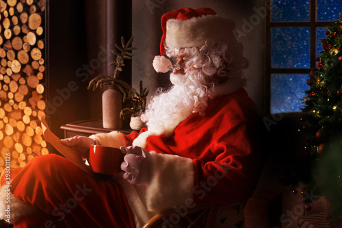 Portrait of happy Santa Claus sitting at his room at home near fireplace and reading Christmas letter or wish list.
