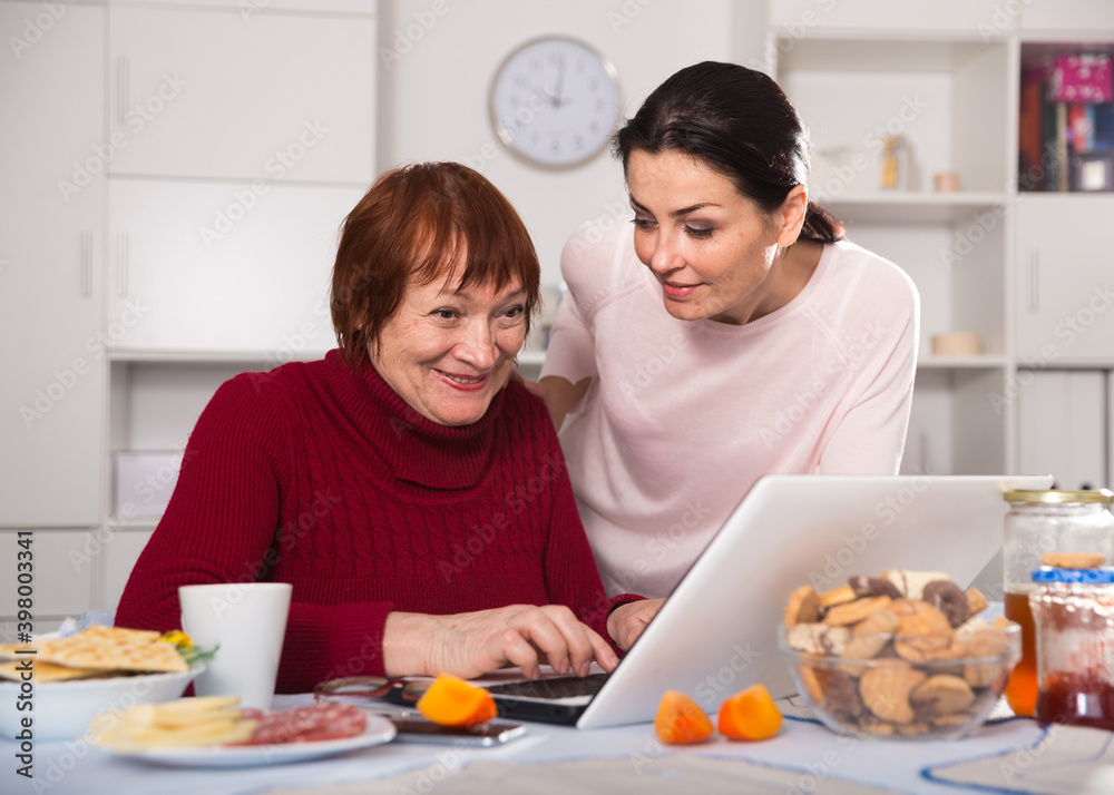 Mature women sitting and using laptop with daughter at table at home.