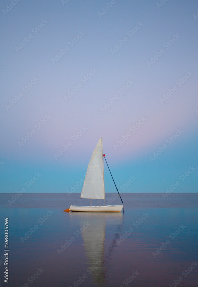 A white boat in the middle of a calmed sea with a pink and blue sunset background. The boat is a sailboat in a peaceful place.
