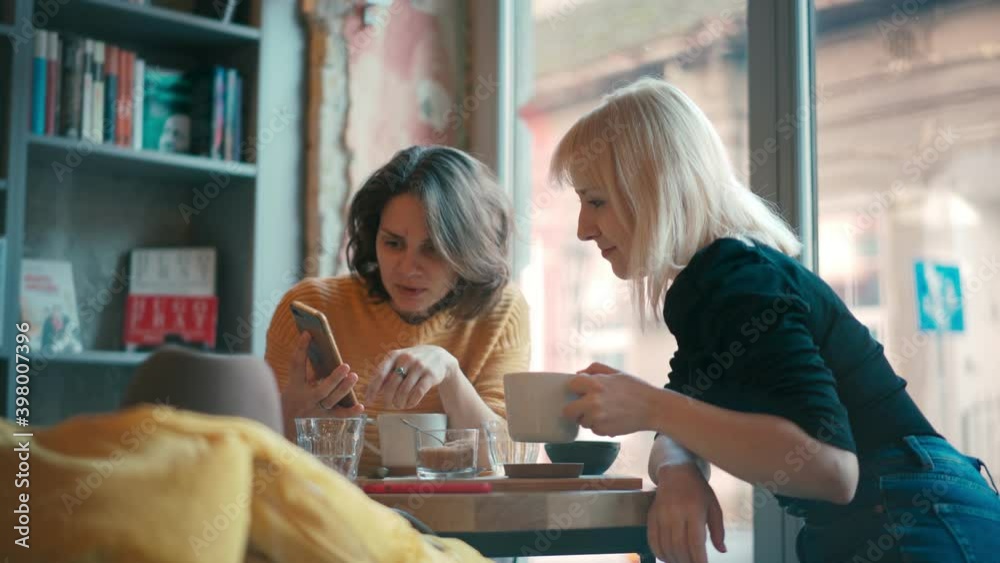 Handheld cinematic shot of two young women sitting at the hipster cafe, having a conversation and showing each other something on their phones.