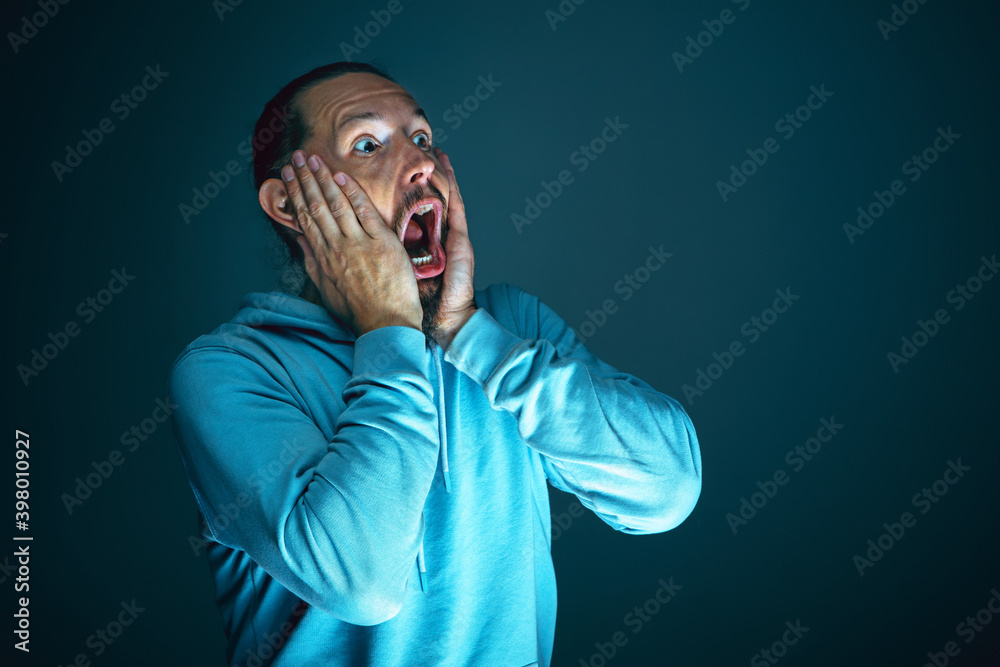 Fototapeta premium Spooky. Portrait of young crazy scared and shocked caucasian man isolated on dark background. Copyspace for ad. Bright facial expression, human emotions concept. Watching horror on TV, cinema.