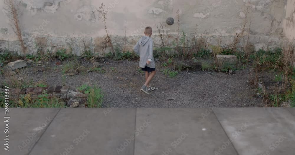 Backside view of tennager boy kicking with feet soccer ball to wall. Kid in hoodie practicing in playing football at abandoned area. Concept of sport and reallife