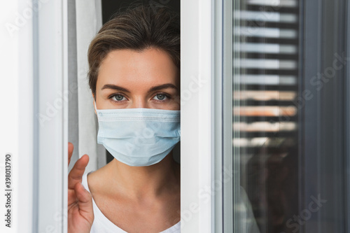 Scared woman wearing prevention mask looking outside through the window