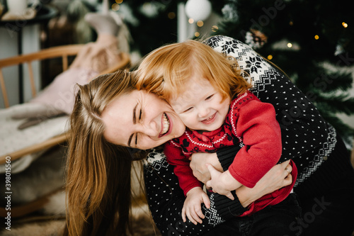 Merry Christmas and Happy Holidays. Cheerful mom hugging cute baby daughter girl near Christmas tree. Mother and little child having fun and playing together at home. Close up.