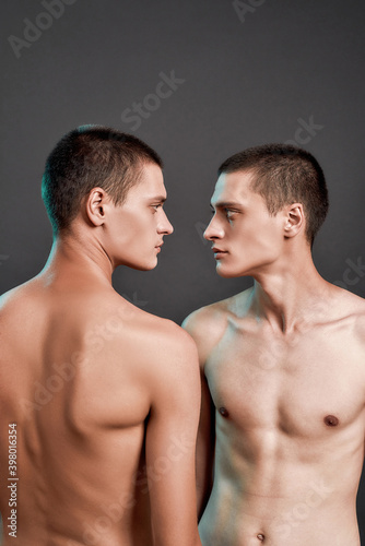 Young caucasian half naked twin brothers looking at each other while posing in studio, standing isolated over grey background