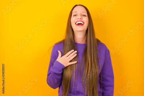 Young beautiful Caucasian woman wearing purple sweater against yellow wall smiles toothily cannot believe eyes expresses good emotions and surprisement