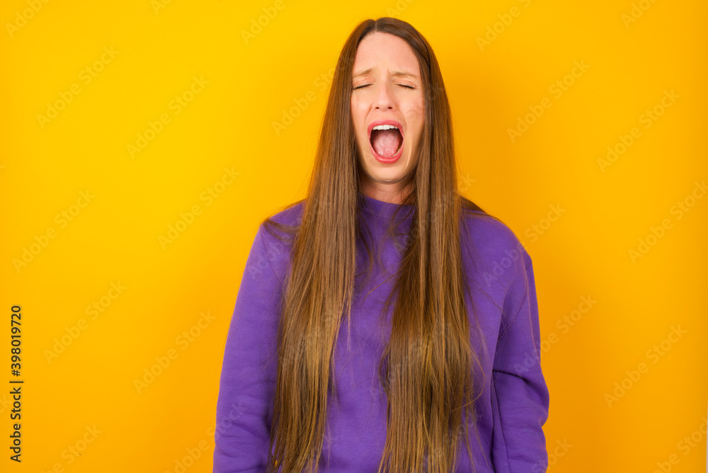 Young beautiful Caucasian woman wearing purple sweater against yellow wall yawns with opened mouth stands. Daily morning routine Young beautiful Caucasian woman wearing purple sweater against yellow wall yawns with opened mouth stands. Daily morning routine