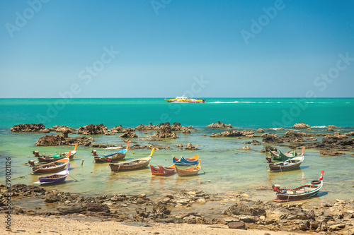 Colorful wooden Thai style fishing boats stand by the rocky coast against the backdrop of the sea with a passenger boat and blue sky
