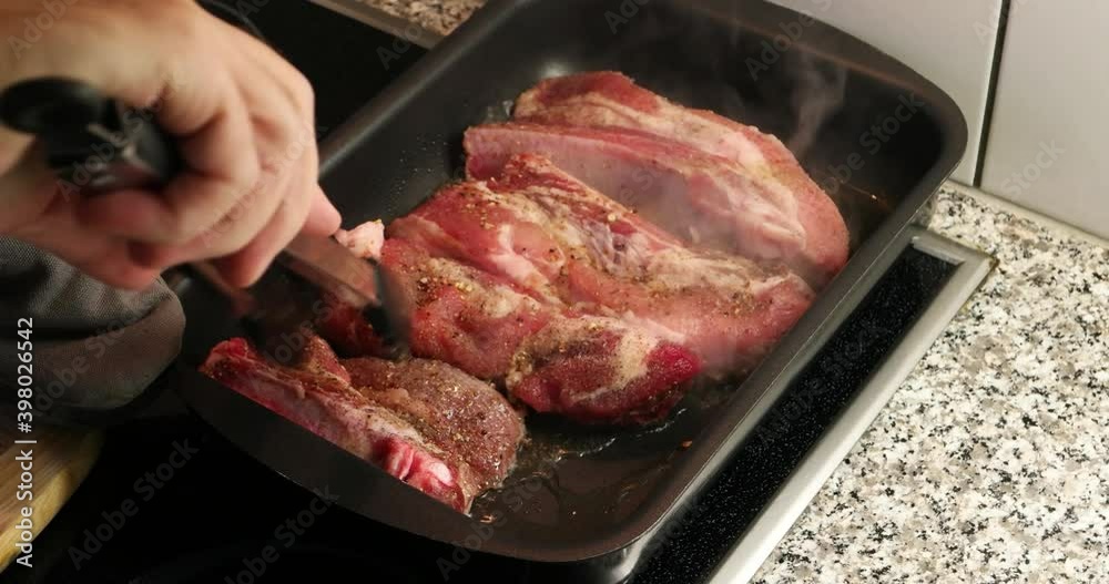 Metal kitchen tongues used to pan sear pork meat with bones in a metal tray on an electric stove. Metal kitchen tongues used to pan sear pork meat with bones in a metal tray on an electric stove.