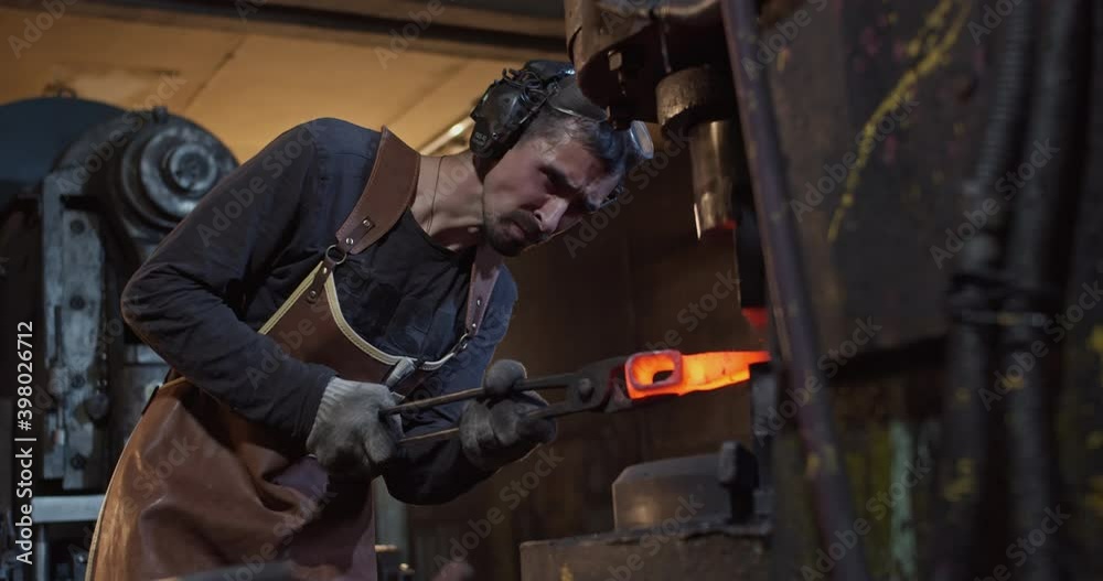 In the workshop, a young blacksmith wearing headphones and a leather apron holds a red-hot workpiece with tongs and shapes it with a mechanical press in slow motion.