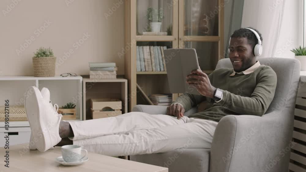 Lockdown of young African man wearing trendy clothes and headphones sitting in armchair with his feet on coffee table, holding tablet in his hands and having online conversation Lockdown of young African man wearing trendy clothes and headphones sitting in armchair with his feet on coffee table, holding tablet in his hands and having online conversation