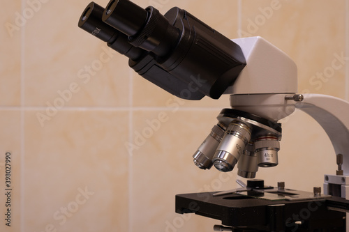 Photo of a light microscope against a tile wall. A gray microscope with a black slide table and black eyepieces. There are several lenses.