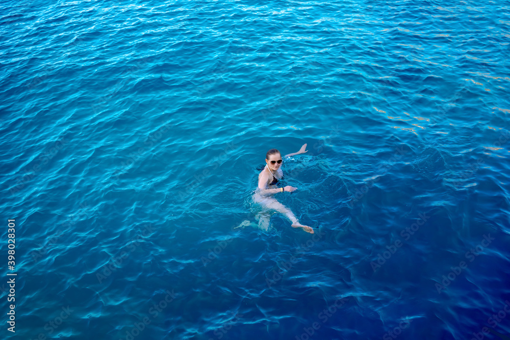 Cute caucasian adult girl in sunglasses swims in blue water - top view. Young woman holds on to the turquoise-blue surface of the sea or pool on a summer sunny day