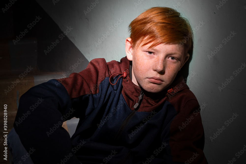 A sad red-haired teenage boy sits alone on the steps in the entrance of an apartment building.