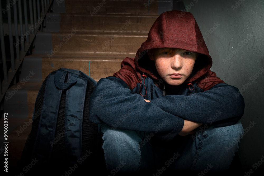 A sad red-haired teenage boy sits alone on the steps in the entrance of an apartment building. A sad red-haired teenage boy sits alone on the steps in the entrance of an apartment building.